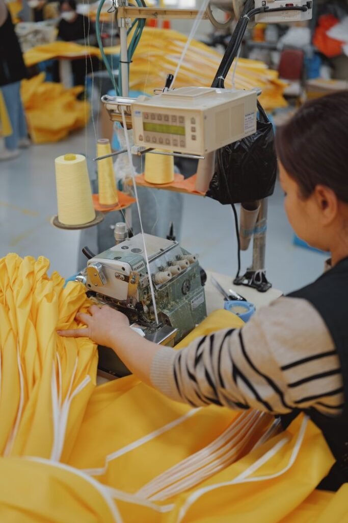 Textile worker using a sewing machine to stitch yellow fabric in a factory setting.