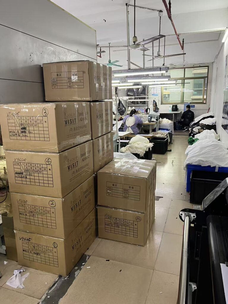 Stacked cardboard cartons beside tables where workers handle white garments
