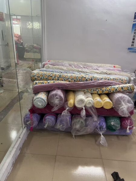 Plastic wrapped fabric rolls in yellow, purple and floral prints stacked beside a glass partition