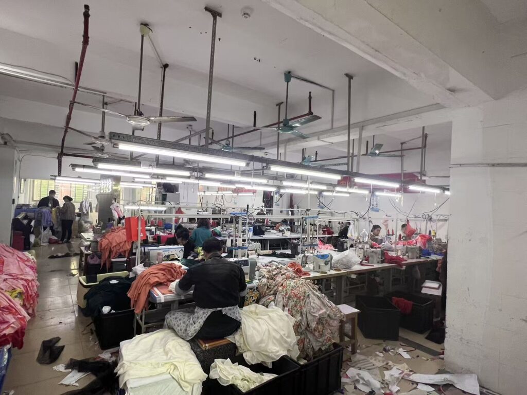 Workers at industrial sewing stations surrounded by fabric and clothing bundles