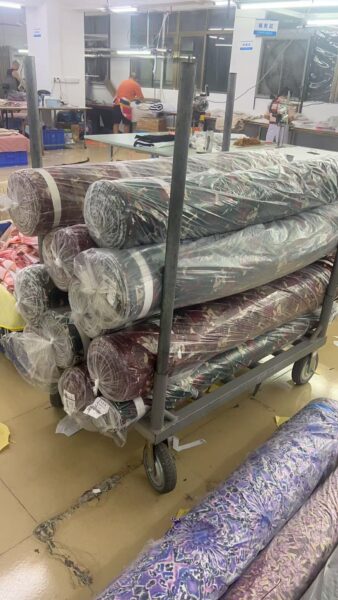 Plastic wrapped printed fabric rolls stacked horizontally on a wheeled trolley