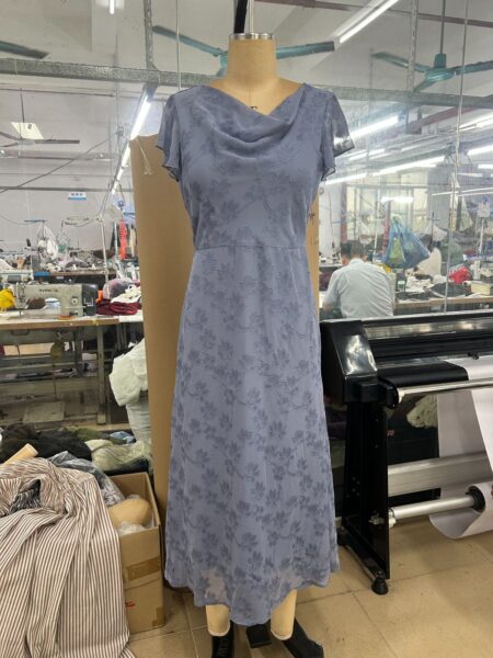 Blue floral cowl-neck midi dress on a mannequin beside a garment workshop