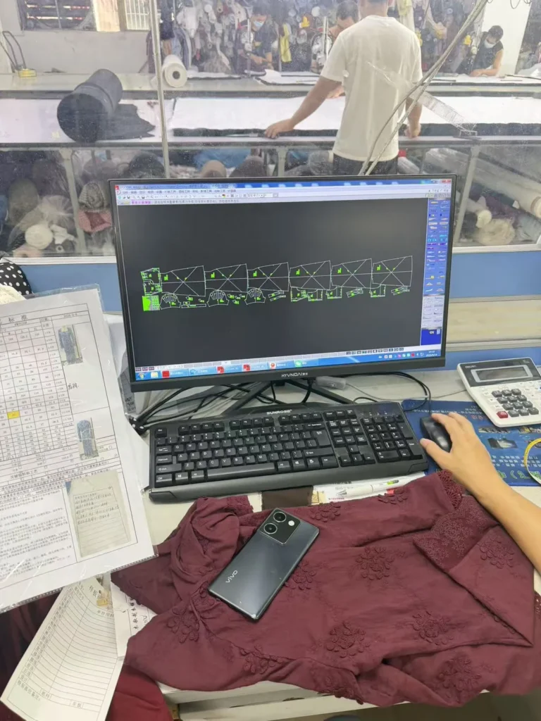 Computer screen displaying garment pattern pieces beside a keyboard and burgundy fabric.